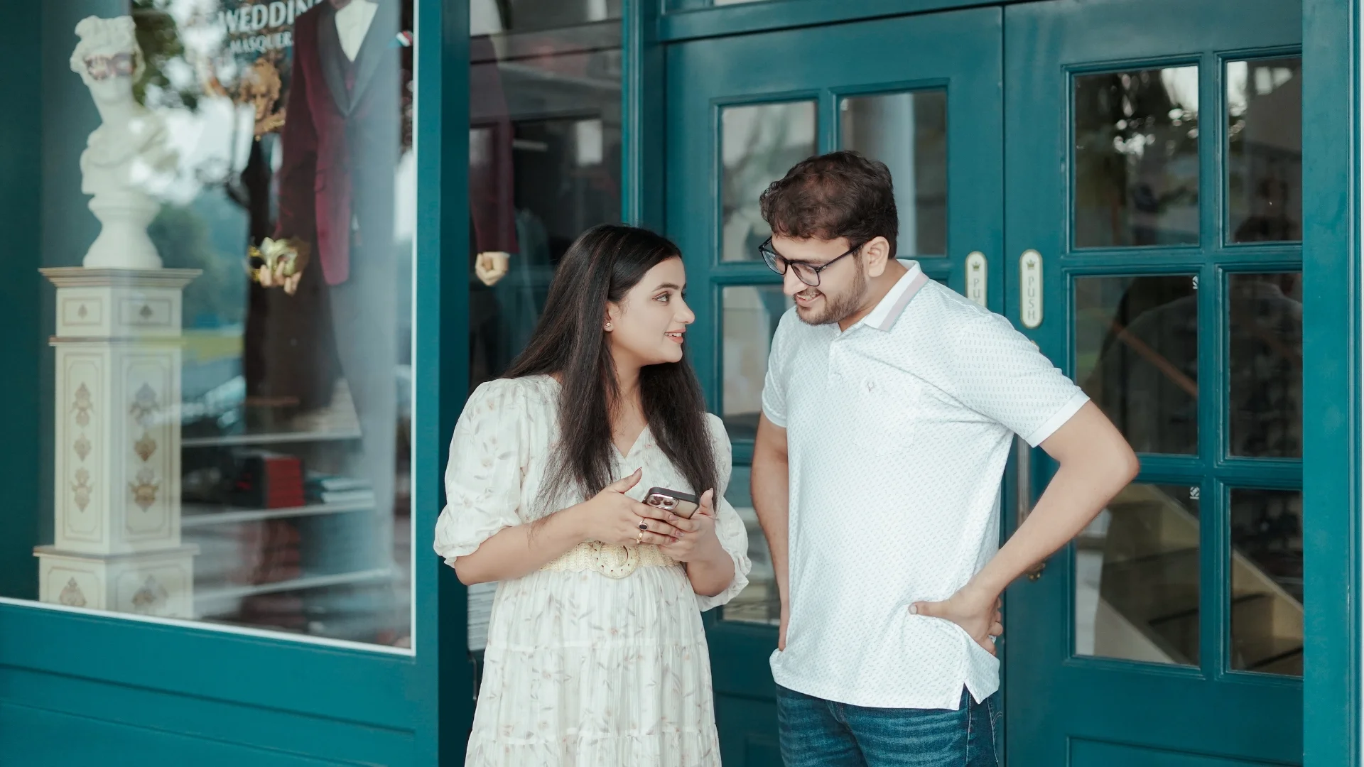 A couple in casual white attire sharing a candid moment while looking at a smartphone outside a vintage teal-colored glass storefront.