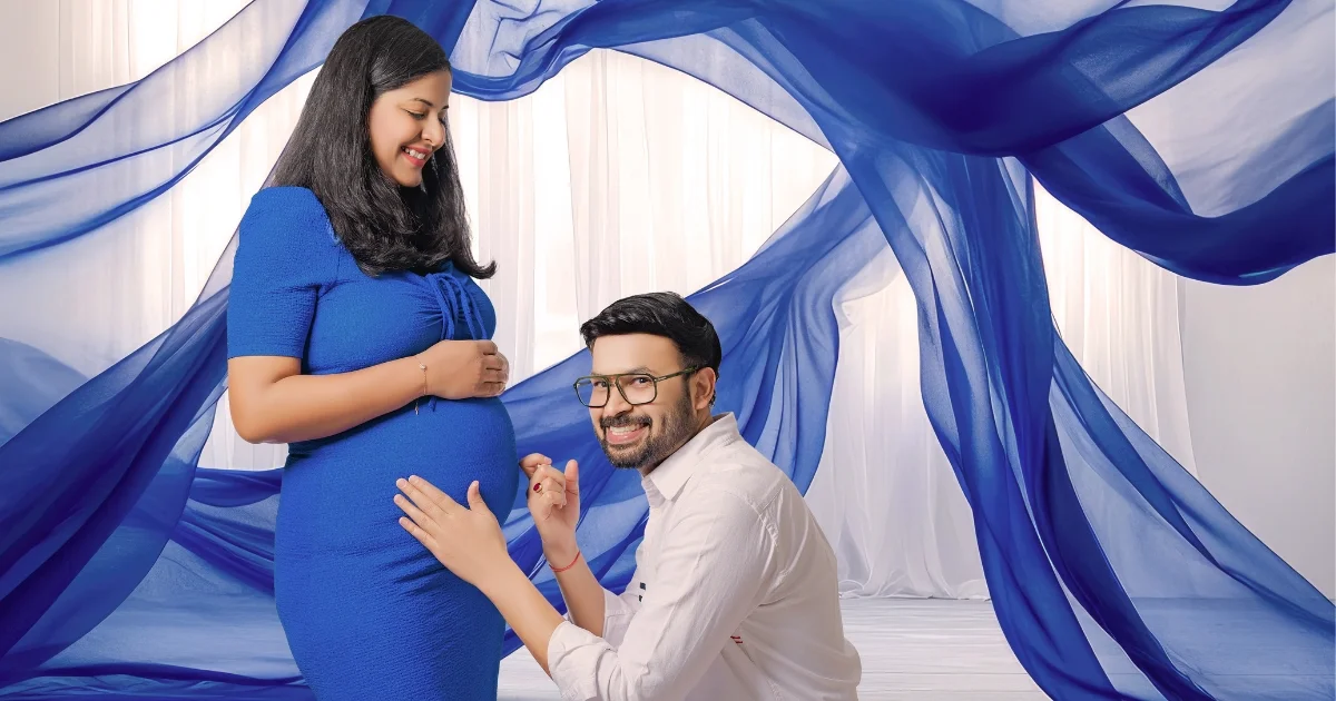 A husband looking at his wife's baby bump against a flowing blue backdrop in an Indo Frames maternity studio session.