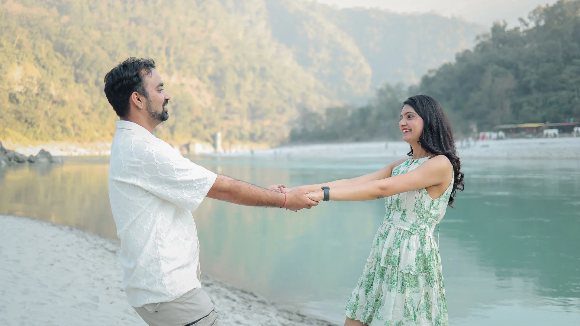 A happy couple leaning back playfully while holding hands on a sandy river beach, captured by Indo Frames photography.