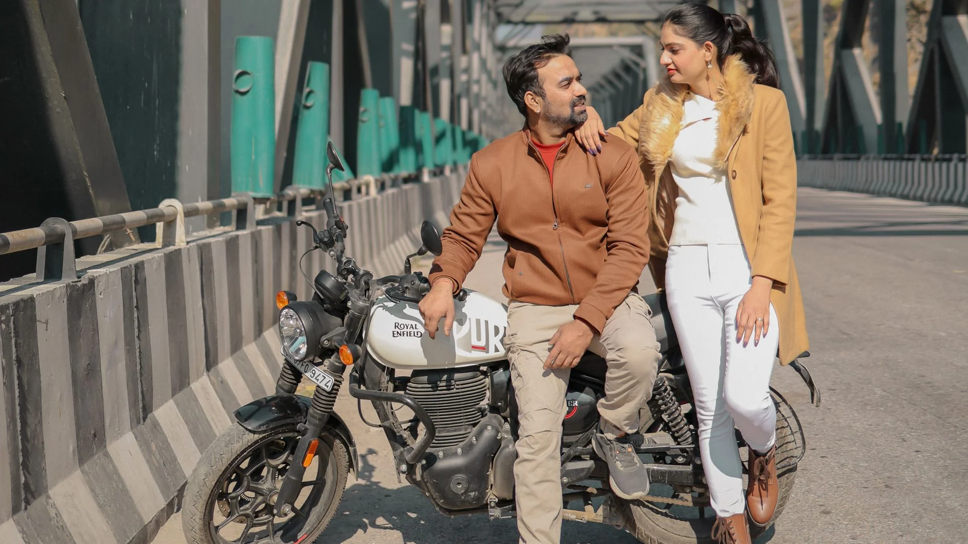 A stylish couple posing with a white Royal Enfield motorcycle on an iron bridge, an outdoor portrait by Indo Frames.