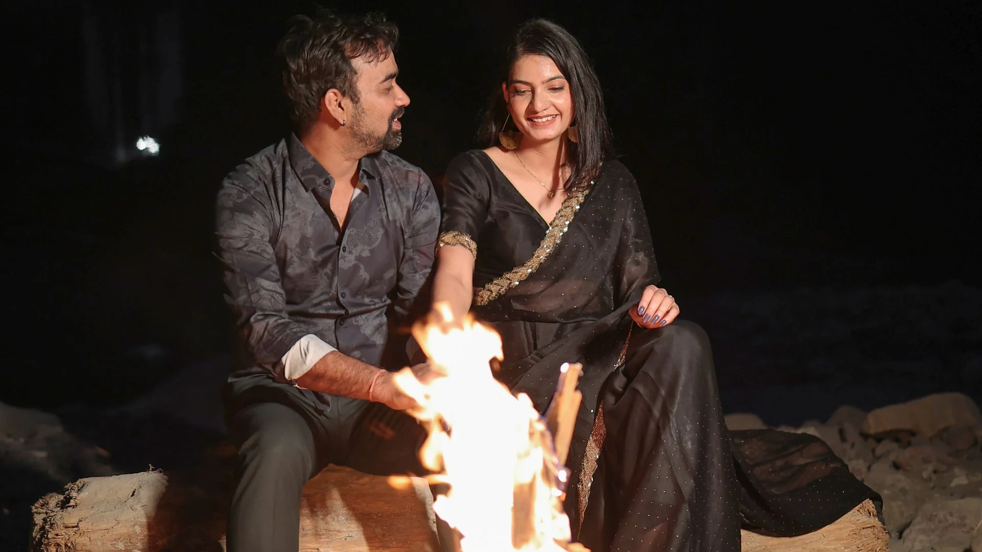 An intimate night photoshoot by Indo Frames featuring a couple sitting by a glowing bonfire, with the woman dressed in a black saree.