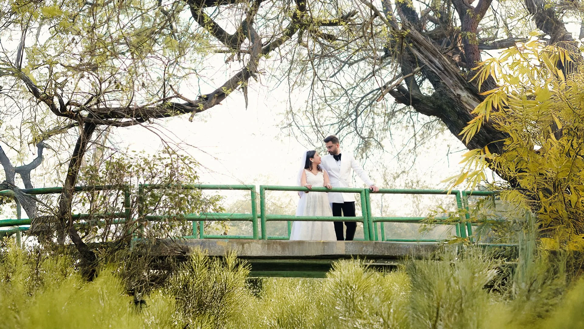A romantic pre-wedding photoshoot by Indo Frames featuring a couple holding hands and walking across a suspension bridge. The woman is in a red gown and the man is wearing a green suit.