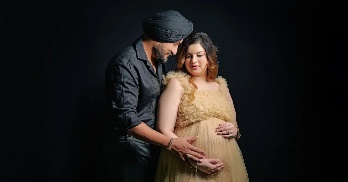 A Sikh couple embracing and holding the pregnancy bump against a black background, a fine art portrait by Indo Frames.