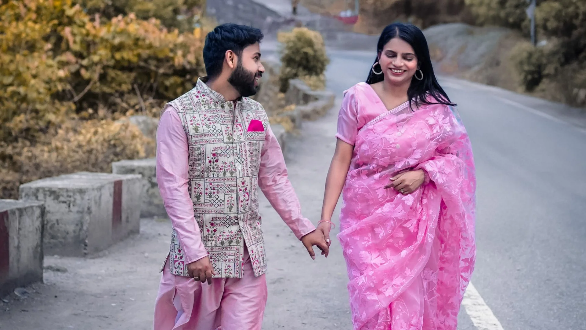 A smiling couple walking along a mountain road in matching pink traditional outfits, shot by Indo Frames.