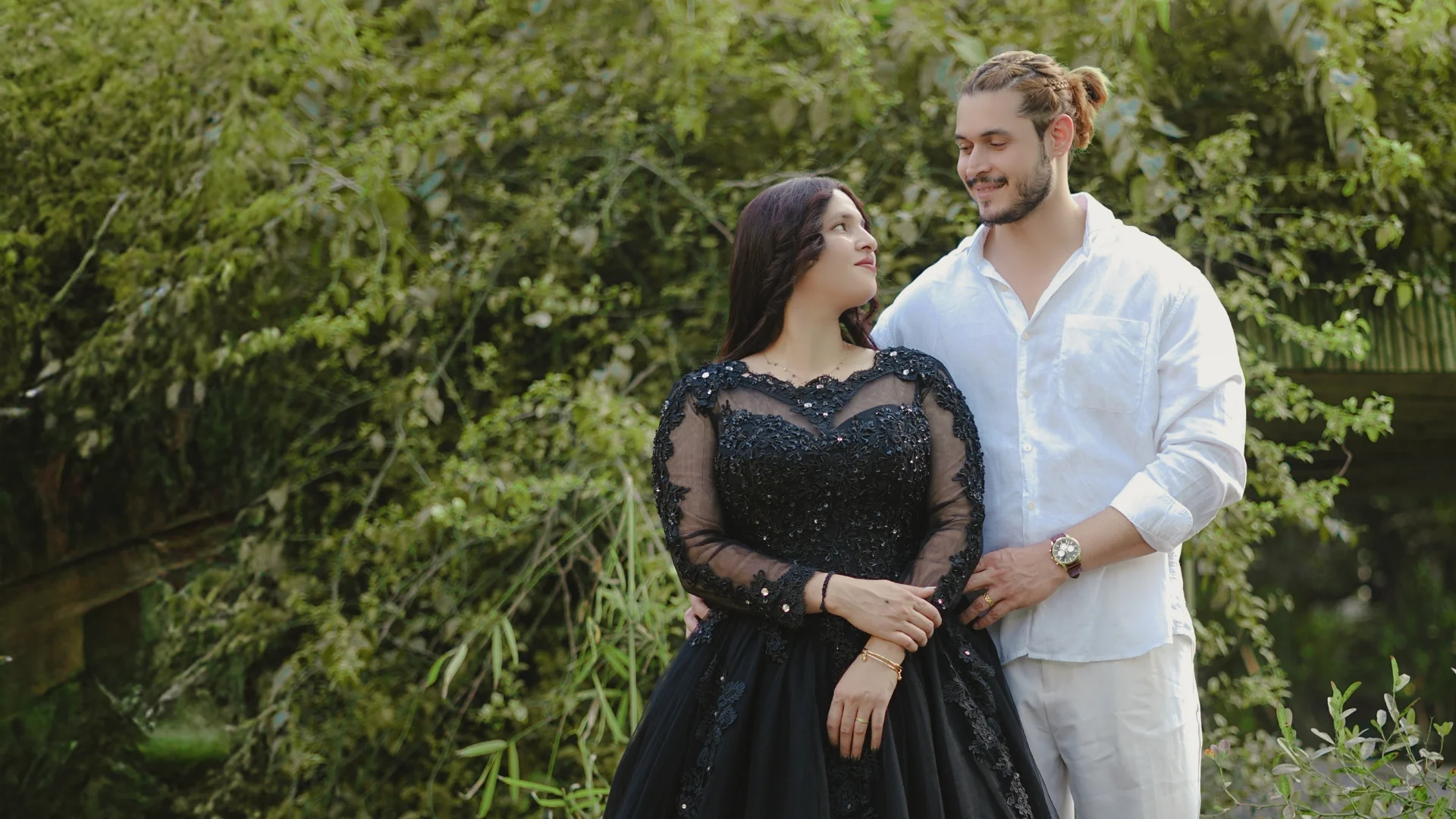 A romantic pre-wedding photoshoot featuring a couple standing outdoors; the woman is wearing an elegant black lace gown and looking up at her partner, who is dressed in a crisp white shirt and matching white trousers against a lush green background.