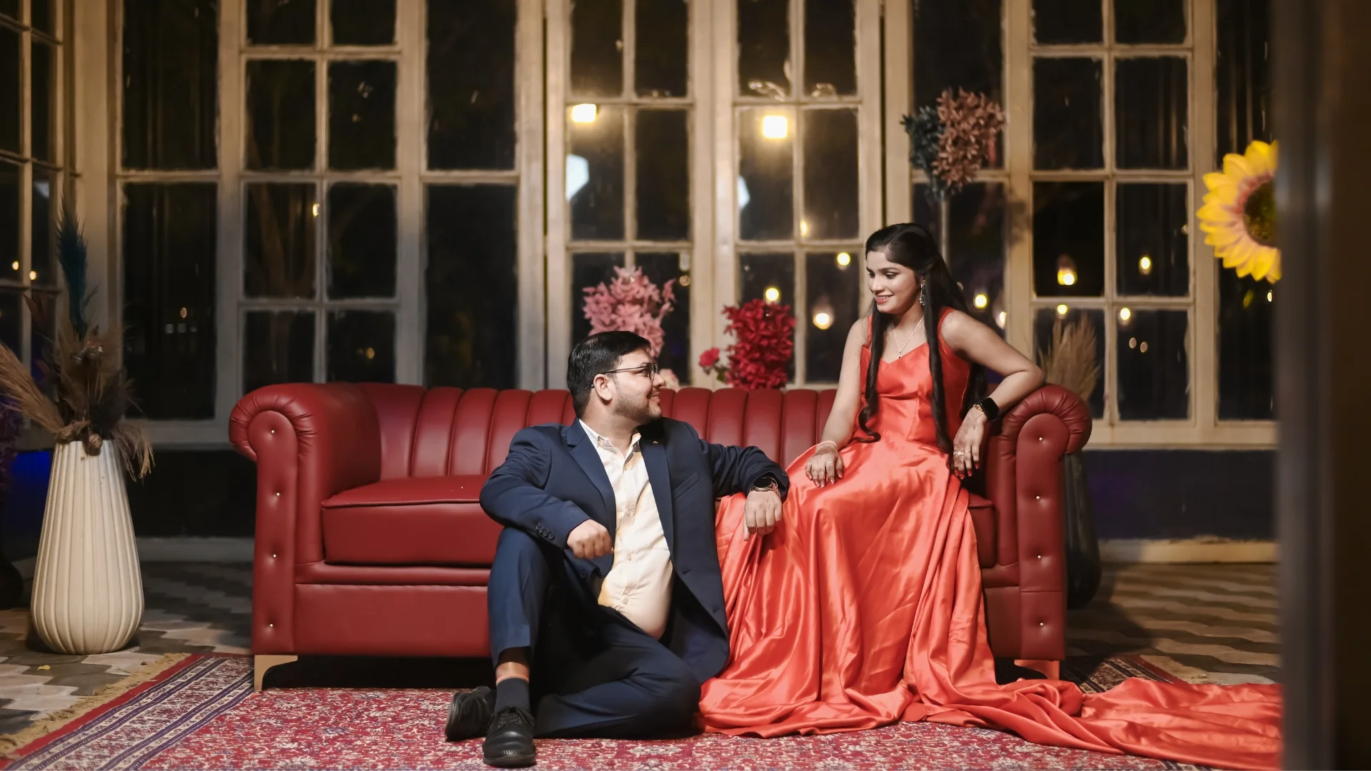 A couple posing in a luxury vintage indoor set, with a woman in a long satin red gown on a chesterfield sofa and a man in a navy blue suit sitting on the rug.